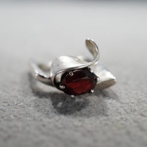 May include: A silver ring with a large, oval, deep red gemstone. The ring has a unique, flowing design with a curved element that wraps around the stone. The ring is set against a gray background.