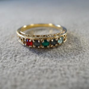 May include: A gold-toned ring featuring a band with an openwork filigree design. The front of the ring is set with five small, round, multi-colored gemstones in a row, including red, dark blue, green, dark blue, and light blue stones.