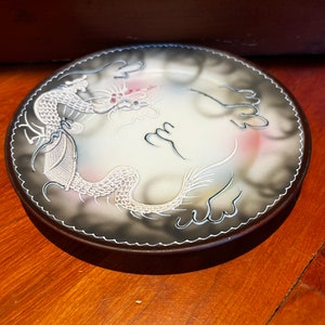 May include: A white ceramic plate with a black rim and a white dragon design. The dragon is surrounded by clouds and has a pink and white color scheme.