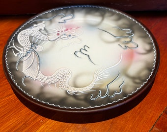 May include: A white ceramic plate with a black rim and a white dragon design. The dragon is surrounded by clouds and has a pink and white color scheme.