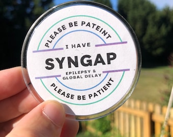 Puede incluir: Una pegatina redonda blanca con el texto "Por favor, sea paciente, tengo SYNGAP, Epilepsia y retraso global, Por favor, sea paciente". La pegatina tiene un borde azul, verde y morado.