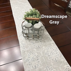 May include: A white table runner with a gray floral pattern. The runner is on a dark wood table with a metal container holding two glass jars. The container has a wooden handle. The text "Dreamscape Gray" is visible in the upper right corner of the image.