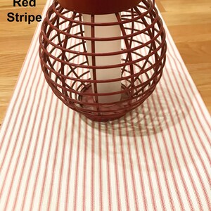 May include: A red metal lantern with a solar panel top sits on a cream-colored table runner with red stripes. The lantern has a cage-like design and a white candle inside. The text "Classic Red Stripe" is in the upper left corner.