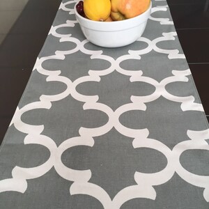 Puede incluir: Un camino de mesa gris con un patrón geométrico blanco. Un tazón blanco con fruta, incluyendo manzanas y limones, está encima del camino de mesa.