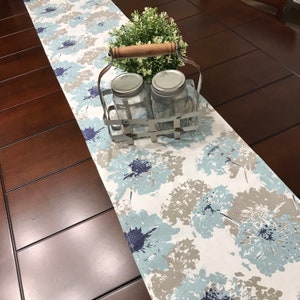 May include: A white table runner with a blue and gray floral pattern. The runner is on a dark brown wooden table with a metal wire basket holding two glass jars and a small potted plant.