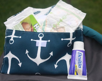 Puede incluir: Una bolsa de tela azul y blanca con estampado de anclas y cierre de velcro blanco. La bolsa está llena de toallitas húmedas para bebés, pañales y un tubo de crema para la dermatitis del pañal Desitin.