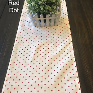 Puede incluir: Un camino de mesa blanco con un patrón de lunares rojos. El camino de mesa está sobre una mesa de madera oscura con una pequeña planta en maceta en la parte superior.