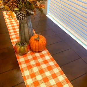 Puede incluir: Un camino de mesa a cuadros naranja y blanco con dos calabazas pequeñas y una jarra de metal con follaje de otoño.