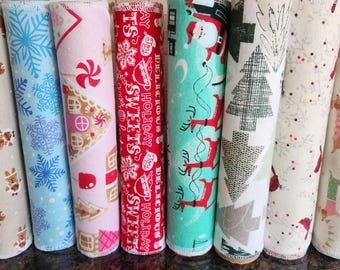 May include: A collection of rolled fabric featuring various holiday-themed patterns. Designs include gingerbread men, snowflakes, candy canes, gingerbread houses, Santa Claus with reindeer, Christmas trees, and dachshunds. The color palette includes red, blue, green, and beige.