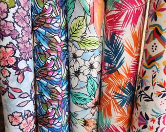 May include: A collection of five rolled fabric cloths with vibrant floral and botanical patterns. The cloths feature a variety of colors, including pink, purple, blue, green, and orange, creating a visually appealing display. The edges are finished with a white trim.