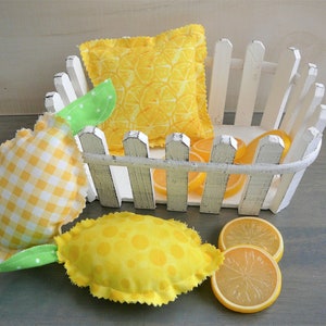 May include: A white picket fence basket holds yellow lemon-themed decorative items. Included are two stuffed lemon-shaped pillows, one with a yellow and white gingham pattern and the other with yellow polka dots. Also, there are several faux lemon slices.