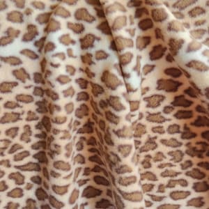 May include: Close-up of a soft, textured fabric with a leopard print pattern. The fabric features a tan background with brown spots of varying sizes and shapes. The material is folded, creating shadows and depth.