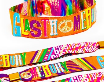 Może przedstawiać: Pomarańczowe materiałowe opaski na nadgarstek z napisem "GLASTONBURY" w kolorowych literach i znakiem pokoju. Opaski mają złote zapięcie i tęczowy sznurek. Opaski są prawdopodobnie przeznaczone na festiwal muzyczny.