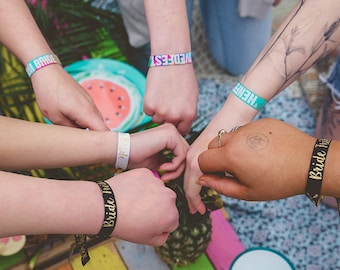 May include: A group of friends wearing colorful wristbands with the text "Bride Tribe" and "Wedfest" on them. The friends are holding hands and smiling.