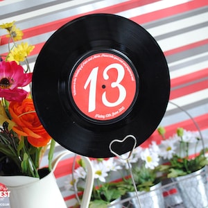 Peut inclure: Un disque vinyle noir avec un centre rouge portant le numéro 13 en blanc. Le disque est maintenu par un porte-carte en fil de fer argenté en forme de cœur. Le texte sur le disque indique "Welcome to the Wedding of Peter and Alison... Friday 12th June 2015."