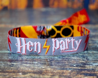 May include: A gray fabric wristband with the embroidered text "Hen Party" in white, accented with a yellow lightning bolt. The interior features a colorful pattern with glasses and other designs. A red and yellow fabric tail is visible.