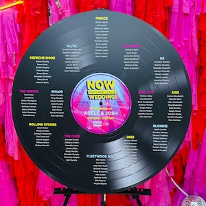 Può includere: Un grande grafico dei posti a sedere per matrimoni in vinile nero con i nomi degli ospiti elencati per artista musicale. Il disco è appoggiato su un supporto nero, con uno sfondo di streamer rosa e rosso.