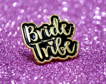 May include: Gold and black enamel pin with the words "Bride Tribe" in a cursive font. The pin has a small heart and arrow design.