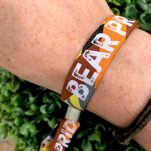 May include: A brown, orange, yellow and grey woven wristband with the text "BEAR PRIDE" printed in white.