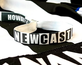 Puede incluir: Una pulsera de tela blanca con la palabra "NEWCASTLE" en letras negras en negrita. Una etiqueta separada dice "HOWAY". La pulsera está sobre un fondo con estampado en blanco y negro.