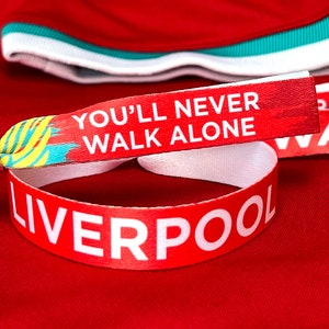 May include: A red fabric wristband with the words "YOU'LL NEVER WALK ALONE" and "LIVERPOOL" in white. The wristband is set against a red background, with a red shirt collar visible.