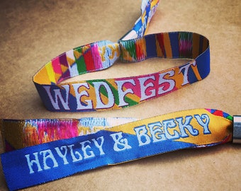 May include: Two colorful wristbands with the text "WEDFEST" and "HAYLEY & BECKY" printed on them. The wristbands are made of woven fabric and have a colorful pattern.