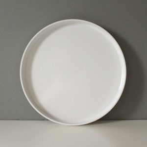 以下が含まれることがあります： わずかに縁が盛り上がった、丸い白いセラミックプレート。プレートは滑らかでマットな仕上がりで、食品の提供に適しています。シンプルなデザインで、さまざまなテーブルセッティングに最適です。