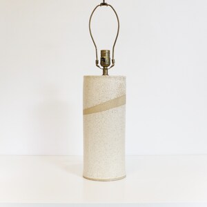 May include: A white ceramic table lamp with a beige stripe. The lamp has a simple, cylindrical design and a brass socket.