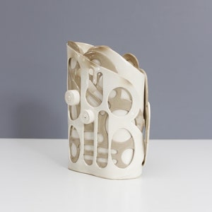 Può includere: Vaso in ceramica bianca con un design a taglio che presenta accenti marroni e forme arrotondate.