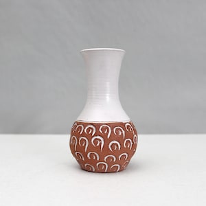May include: A small, handmade ceramic vase with a white upper section and a textured, reddish-brown lower section. The lower section features a repeating pattern of white, curved lines. The vase has a narrow neck and a rounded base, suitable for displaying small flowers.