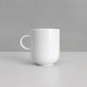 Puede incluir: Una taza de cerámica blanca lisa con un cuerpo cilíndrico y una base redondeada. La taza presenta un asa sencilla y curva en el lado izquierdo, ofreciendo una apariencia limpia y uniforme sin patrones, texturas ni elementos decorativos.
