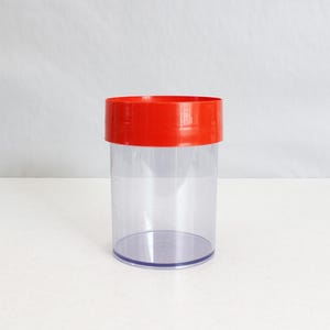 May include: A clear plastic container with a bright red lid. The cylindrical container is transparent, allowing visibility of its contents. The lid sits snugly on top, creating a secure seal. The container is designed for storage.