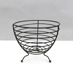 May include: A black wire fruit basket with a rounded bowl shape. The basket has a series of horizontal wire rings held together by vertical supports. It stands on three small, curved legs with rounded tips. The basket is empty.