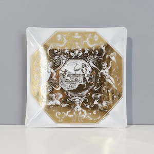 Pode incluir: Um cinzeiro quadrado de vidro branco com um design octogonal dourado que apresenta um querubim com um arco e flecha. O design é rodeado por ornamentos florais e de volutas.