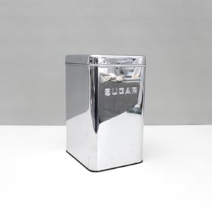 May include: A rectangular, polished chrome sugar canister with rounded corners and a matching lid. The word SUGAR is embossed in capital letters on the front surface of the container, which features a highly reflective, mirror-like metallic finish.
