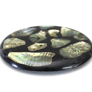 Può includere: Un oggetto decorativo rotondo e nero con intarsi di pezzi di conchiglia di abalone iridescenti. I frammenti di conchiglia, di varie dimensioni e forme, mostrano sfumature di verde, argento e oro. L'oggetto ha una finitura lucida.