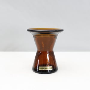 Può includere: Un portacandele in vetro marrone a forma di clessidra. Ha una parte superiore e inferiore ampia, con una sezione centrale più stretta. Il vetro è di un ricco colore ambra e il pezzo ha una finitura lucida. L'etichetta recita "DANSK DESIGNS LTD."