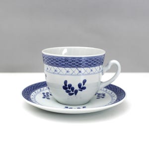 Puede incluir: Juego de taza y platillo blancos con diseños florales y geométricos azules. La taza tiene un asa y una banda con diseños azules en la parte superior. El platillo tiene un diseño a juego. El juego está sobre una superficie blanca.