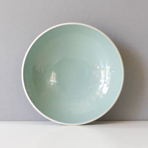 May include: A round, pale blue ceramic bowl with a white rim. The bowl has a smooth, glossy finish. It is set against a grey background. Suitable for serving salads, soups, or other dishes. Measurements are in centimetres.
