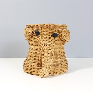 Könnte beinhalten: Ein geflochtener Korb aus Weide, der wie ein Elefant geformt ist, mit schwarzen Knopfaugen. Der Korb ist hellbraun und hat eine natürliche geflochtene Textur.