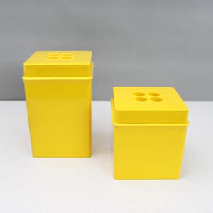 May include: Two bright yellow, rectangular containers with square lids. Each lid has four circular openings. The containers are set against a neutral grey background.