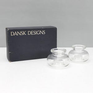 Puede incluir: Dos candelabros de vidrio transparente con forma redonda y una pequeña abertura. Una caja negra con el texto "DANSK DESIGNS" está detrás de los candelabros. Los candelabros están sobre una superficie blanca.