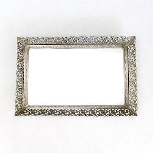 May include: A rectangular vanity mirror with an ornate, silver-colored filigree frame. The mirror's surface is clear and reflective, and the frame features a detailed, repeating floral pattern. The mirror is set against a plain white background.