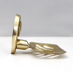 May include: A brass soap dish with a square, slotted holder and a circular backplate. The dish is angled, showcasing its design and the warm, metallic tones of the brass. The soap dish is designed to hold a bar of soap.