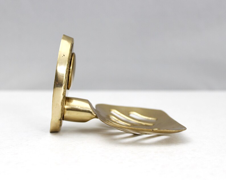 May include: A brass soap dish with a square, slotted holder and a circular backplate. The dish is angled, showcasing its design and the warm, metallic tones of the brass. The soap dish is designed to hold a bar of soap.