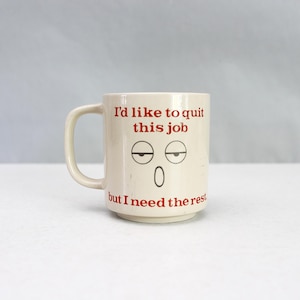 May include: A cream-coloured ceramic mug with a handle. The mug features the text "I'd like to quit this job" in red, along with a face graphic. Below the text is the phrase "but I need the rest."