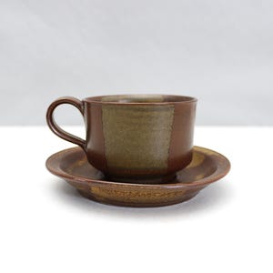 May include: A ceramic teacup and saucer set. The cup and saucer are brown with a rectangular olive green accent on the cup. The set is on a white surface.