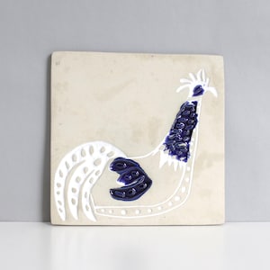 May include: A white ceramic tile with a blue and white rooster design. The rooster is stylized with a long neck and tail feathers. The tile is a decorative piece for a kitchen or bathroom.