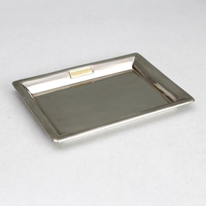 Op de afbeelding: Een rechthoekig dienblad van zilverkleurig metaal met een glad, reflecterend oppervlak en een opstaande rand. Een klein, rechthoekig accentplaatje van goudkleurig metaal is in het midden van de bovenrand geplaatst voor een subtiel contrast.