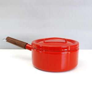 May include: A red enamel saucepan with a lid and a wooden handle. The saucepan is round and has a shiny finish. The handle is attached to the side of the pan with a metal bracket.